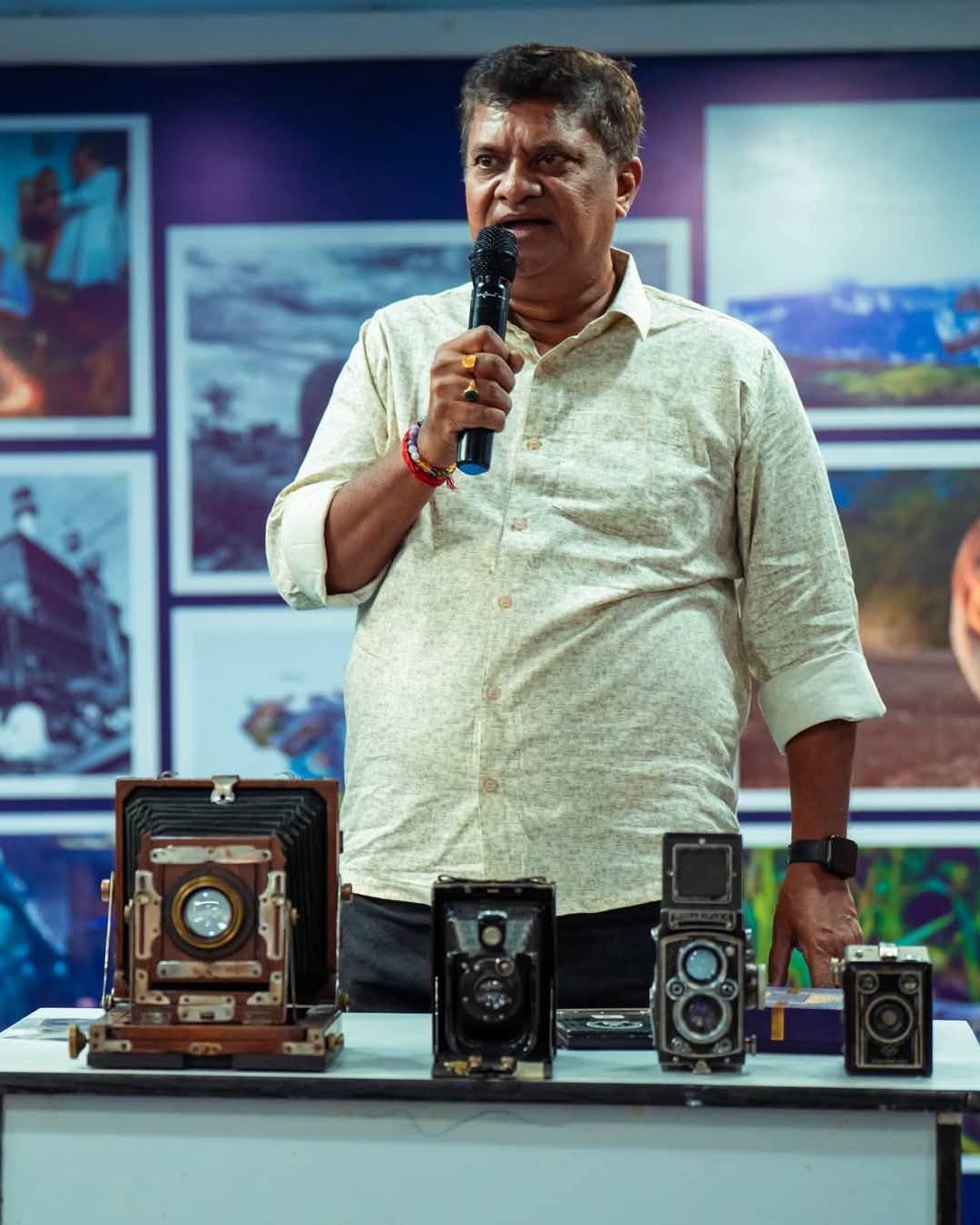 Jayanta Shaw speaking into a microphone at an exhibition, standing behind a table of vintage cameras — a wooden large-format field camera, two twin-lens reflexes and a box camera — with framed photographs on the wall behind him.